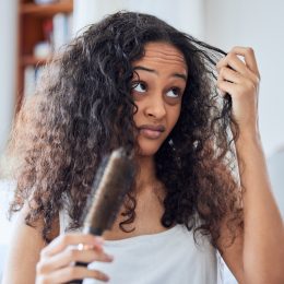 Woman in a white tank top in her bedroom trying to brush her thick, curly hair
