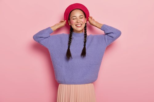 Young woman smiling in braided pigtails, wearing a pink hat, purple sweater, and peach skirt, against a pink background