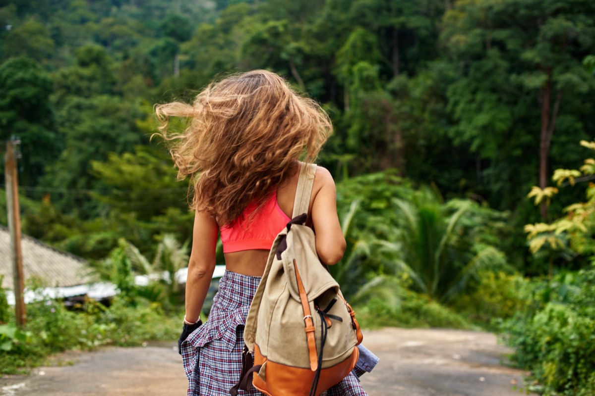 Millenial young woman in pink top bra with backpack flapping with her curly hair is hiking in mountains through wet green jungle. Actively spending summer vacation Active hobby