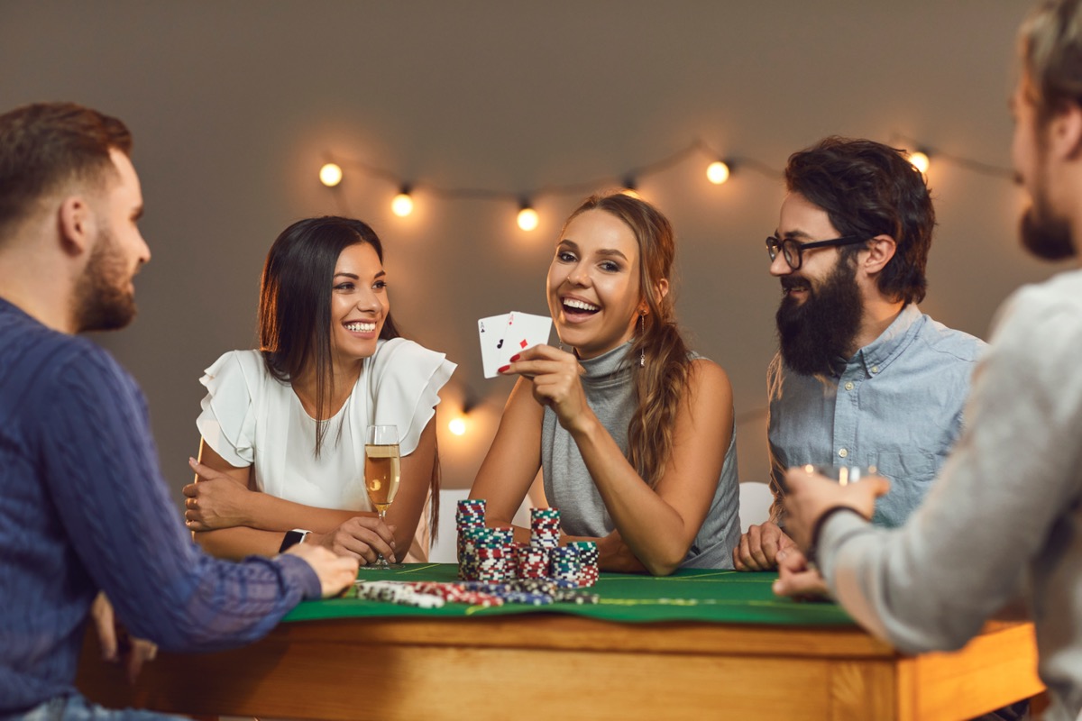 Happy young woman showing two aces while playing poker while sitting at a game table with her friends. Concept of themed celebrations of birthdays or corporate events in a casino or at home.