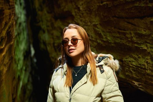 A young woman hiking in a cave wearing a light green down jacket, black shirt, and sunglasses 