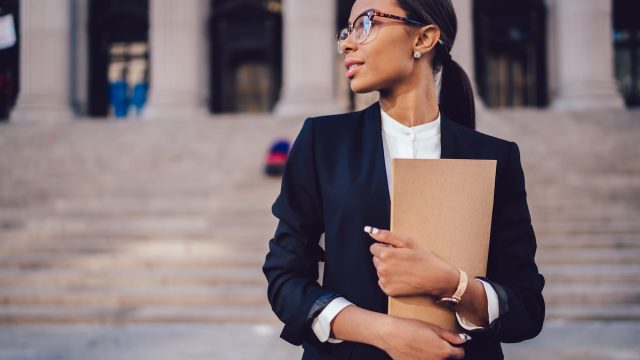 Pensive African American female lawyer in stylish formal suit holding folder with mock up area and looking away standing against courthouse. Half length of woman professional advocate with documents