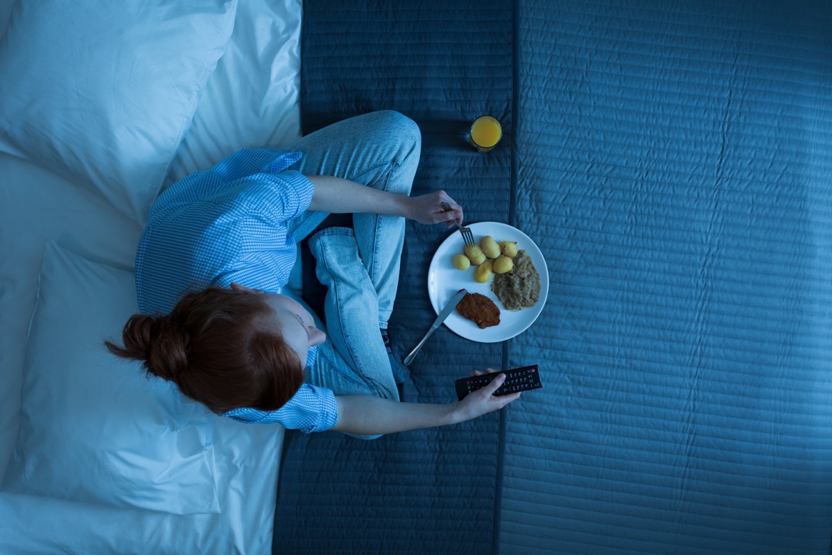 Top view of a woman sitting on a bed, eating dinner and holding a remote control