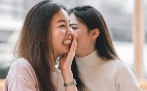 Woman whispering funny paranoia questions into another woman's ear