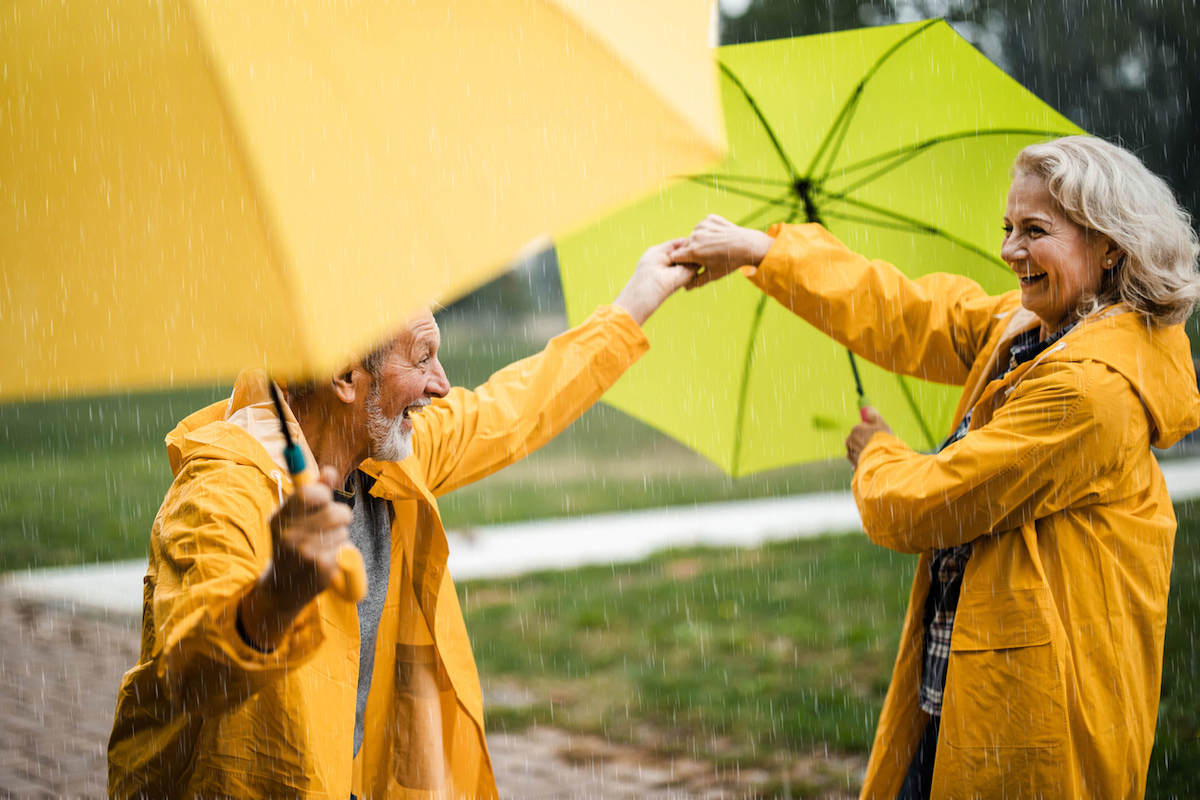 Happy senior couple in raincoats having fun while dancing with umbrellas during rainy day at the park.