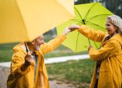 Happy senior couple in raincoats having fun while dancing with umbrellas during rainy day at the park.