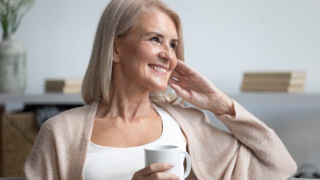 A happy mature woman sitting on her couch with a cup of coffee, gazing to the side.