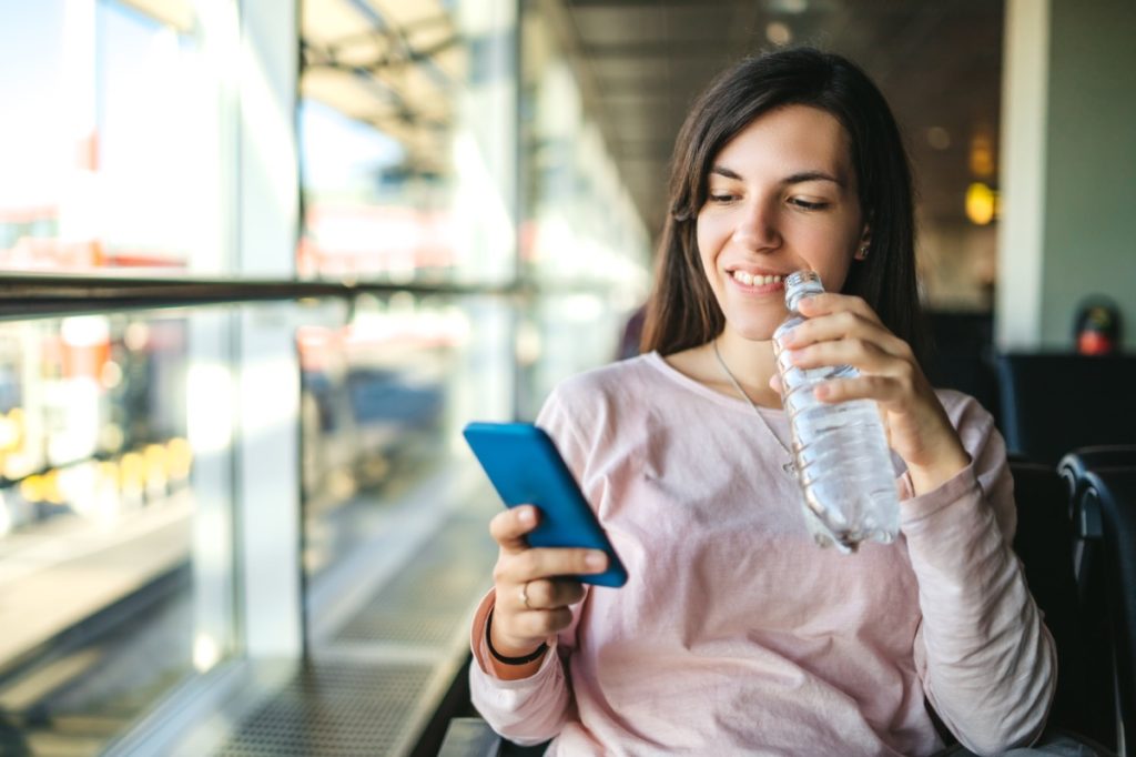 Young woman drinking bottled water and using phone at the airport departure area