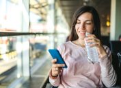 Young woman is drinking bottled water and using phone at the airport departure area