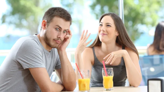 Man looking away and rolling his eyes at a talkative woman while at a restaurant table having orange juice.