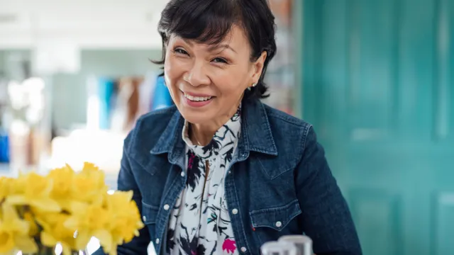Mature woman smiling wearing a jean jacket and a blouse next to a vase of daffodils