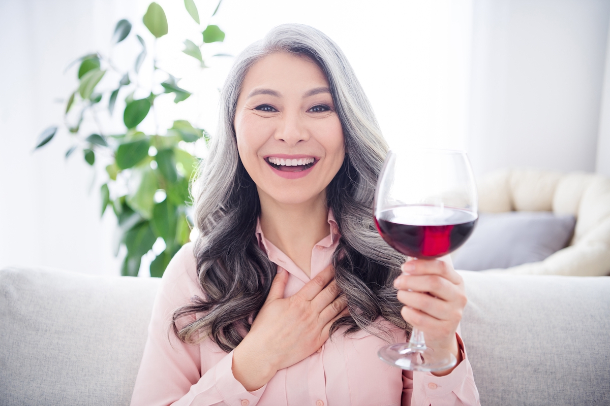 Portrait of attractive cheerful gray-haired woman sitting on divan drinking wine