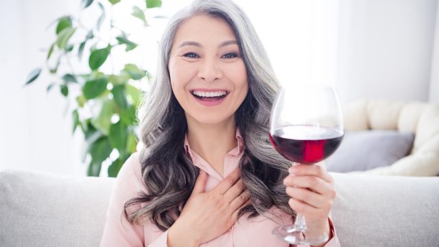 Portrait of attractive cheerful gray-haired woman sitting on divan drinking wine