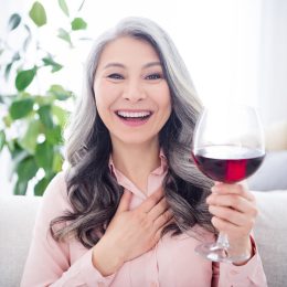 Portrait of attractive cheerful gray-haired woman sitting on divan drinking wine