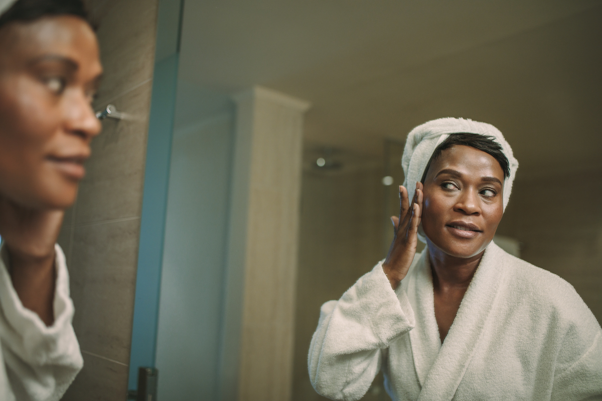 A mature woman wearing a white bathrobe and white hair towel stands in front of the mirror touching her face.
