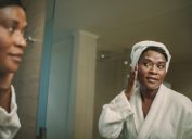 A mature woman wearing a white bathrobe and white hair towel stands in front of the mirror touching her face.