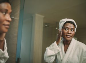 A mature woman wearing a white bathrobe and white hair towel stands in front of the mirror touching her face.