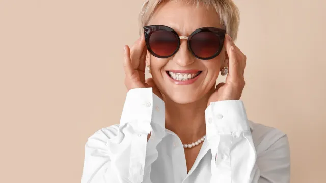 A stylish older woman wearing a white shirt, pearl jewelry, and large black sunglasses against a beige background