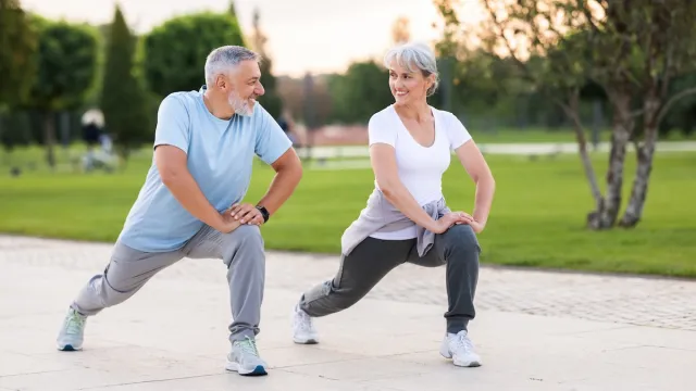 A middle-aged man and woman, both with gray hair, exercise outside. They're stretching doing lunges.