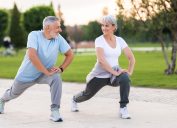 A middle-aged man and woman, both with gray hair, exercise outside. They're stretching doing lunges.