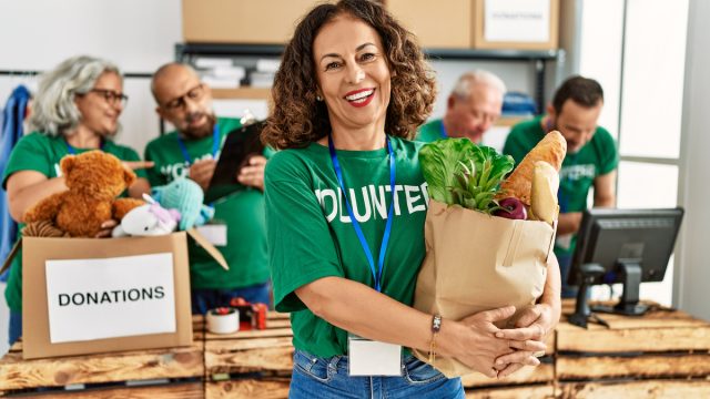 A smiling middle-aged woman wearing a green "volunteer" t-shirt holds a bag of food to be donated.