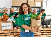 A smiling middle-aged woman wearing a green "volunteer" t-shirt holds a bag of food to be donated.
