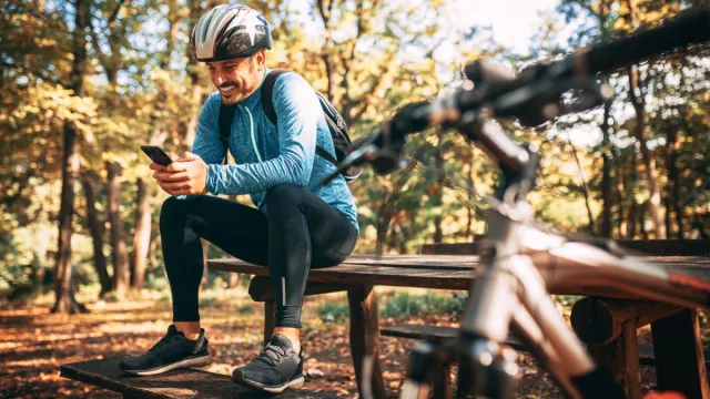 male cyclist sitting on a bench in the forest playing offline games on his phone