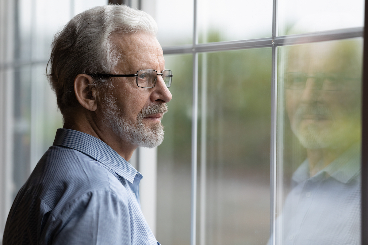 A pensive older man wearing a button-down shirt and glasses with gray hair is looking out a window
