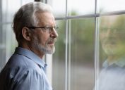 A pensive older man wearing a button-down shirt and glasses with gray hair is looking out a window
