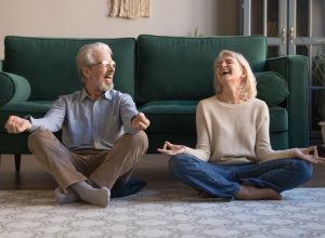 Happy mature couple having fun, practicing yoga together at home, laughing grey haired man and woman sitting in lotus pose on floor in living room, breathing, relaxing, healthy lifestyle concept