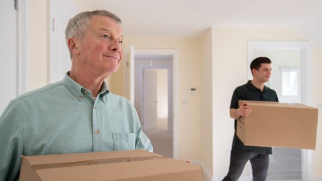 Senior Man Downsizing In Retirement Carrying Boxes Into New Home On Moving Day With Removal Man Helping