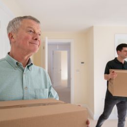 Senior Man Downsizing In Retirement Carrying Boxes Into New Home On Moving Day With Removal Man Helping