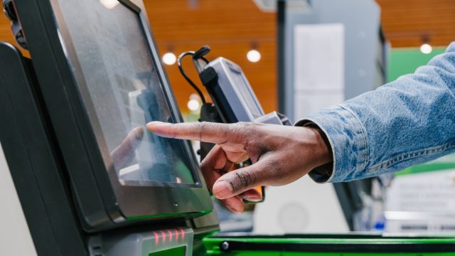 man finger touches screen of self-pay point to pay in shopping mall supermarket light hall extreme close view