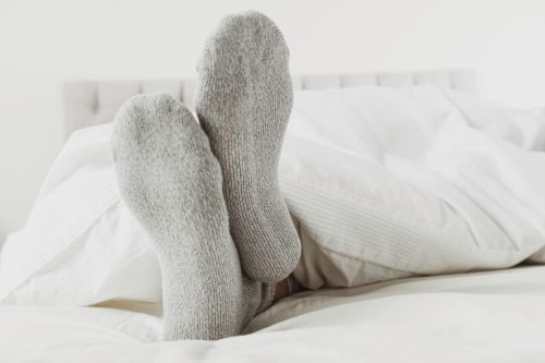 Close up of feet wearing gray socks in bed with white sheet.