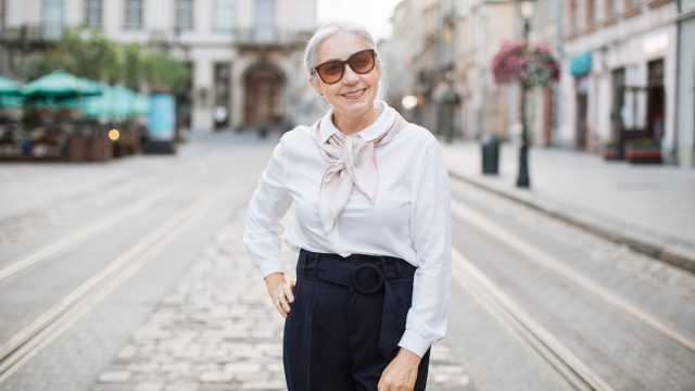 Adult grey-haired woman in elegant outfit and trendy sunglasses posing and smiling outdoors on an old city street.
