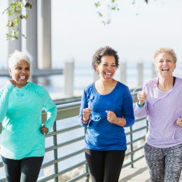 A group of three multi-ethnic women in their 50s and 60s power walking on a city waterfront side by side. They are having a good time, laughing.