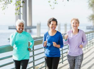 A group of three multi-ethnic women in their 50s and 60s power walking on a city waterfront side by side. They are having a good time, laughing.