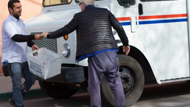 A senior man hands a letter to a smiling postal worker on a street in downtown Santa Fe. Close-up shot with a mail truck also in the frame.