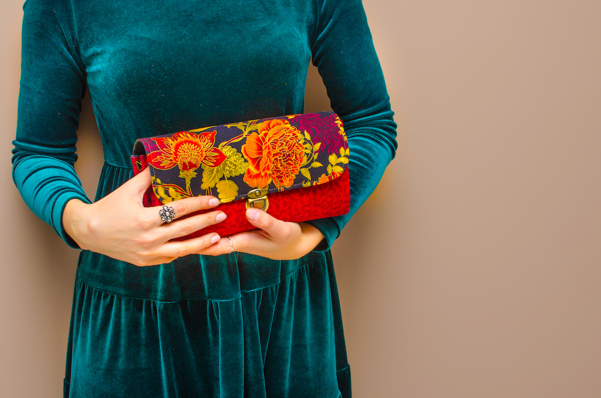 In the hands of a young woman wearing an elegant green blue velvet dress, a bright red clutch handbag with floral print on brown background. Women's day, fashion, spring, flower, rose, romance, love.