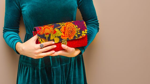 In the hands of a young woman wearing an elegant green blue velvet dress, a bright red clutch handbag with floral print on brown background. Women's day, fashion, spring, flower, rose, romance, love.