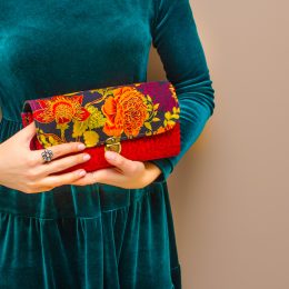 In the hands of a young woman wearing an elegant green blue velvet dress, a bright red clutch handbag with floral print on brown background. Women's day, fashion, spring, flower, rose, romance, love.