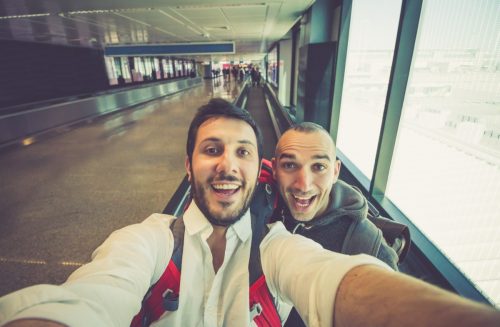 Two men taking a selfie together on a moving walkway at the airport