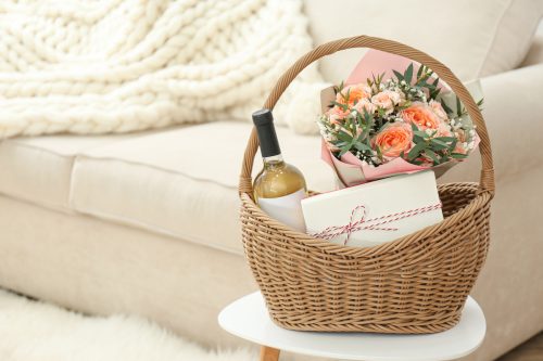 A wicker gift basket filled with pink roses, wine, and a pastry box sits on a stool in front of a cream-colored couch