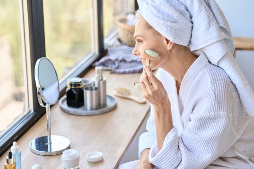A mature woman in a bathrobe and towel sitting in front of window using a jade roller on her face in the mirror 