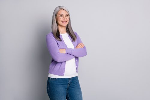 A mature woman with shoulder-length gray hair wearing a white t-shirt, blue jeans, and purple cardigan stands against a gray background