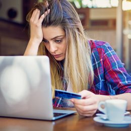A young woman sitting at her laptop while holding her credit card with a stressed look on her face