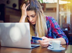 A young woman sitting at her laptop while holding her credit card with a stressed look on her face