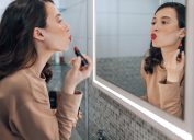A brunette woman looking in the mirror puckering her lips as she applies red lipstick