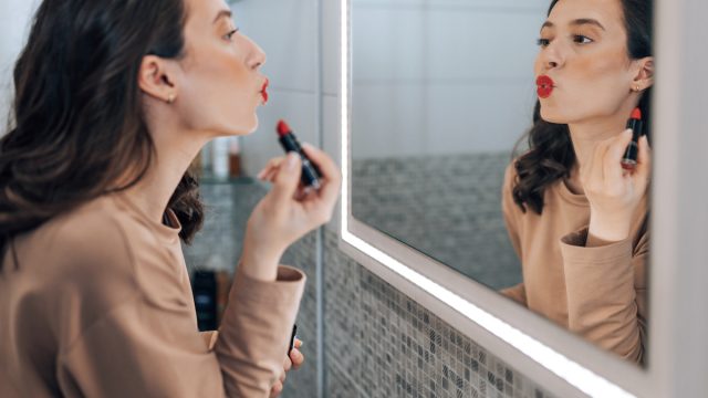 A brunette woman looking in the mirror puckering her lips as she applies red lipstick