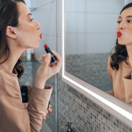 A brunette woman looking in the mirror puckering her lips as she applies red lipstick
